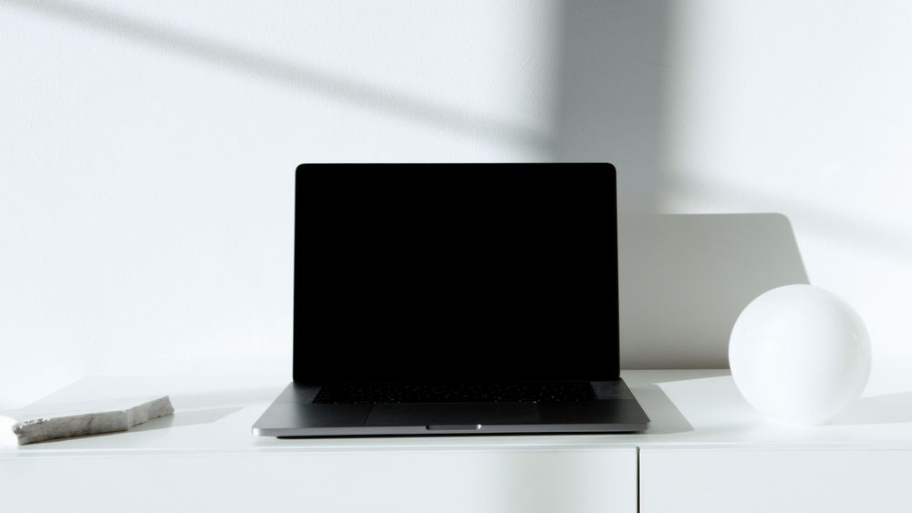 Escritorio blanco minimalista con una notebook cerrada y luz lateral, en blanco y negro
