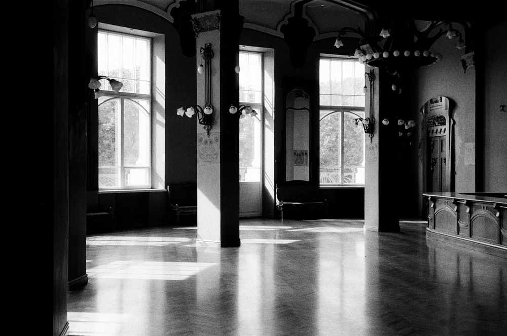 Salón antiguo vacío con ventanales altos, columnas y piso de madera iluminado por el sol, en blanco y negro